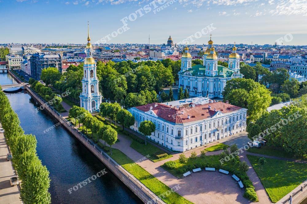 На переднем плане изображены Николо-Богоявленский морской собор с его великолепными золотыми куполами и высокая колокольня, расположенные у канала Грибоедова. Собор окружен зелеными деревьями, создающими уютную летнюю атмосферу. Справа можно увидеть часть исторического здания, гармонирующего с окружающим пейзажем. На заднем плане виднеется Исаакиевский собор и другие достопримечательности Санкт-Петербурга, подчеркивающие величие города.